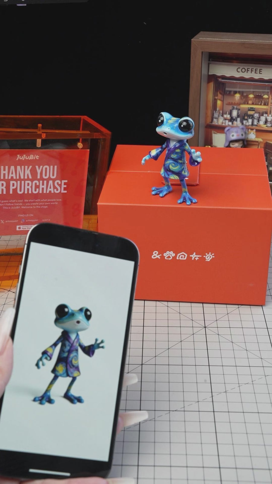 Custom frog figurines in different colors compared to their phone photo references beside a JuJuBit box - JuJuBit